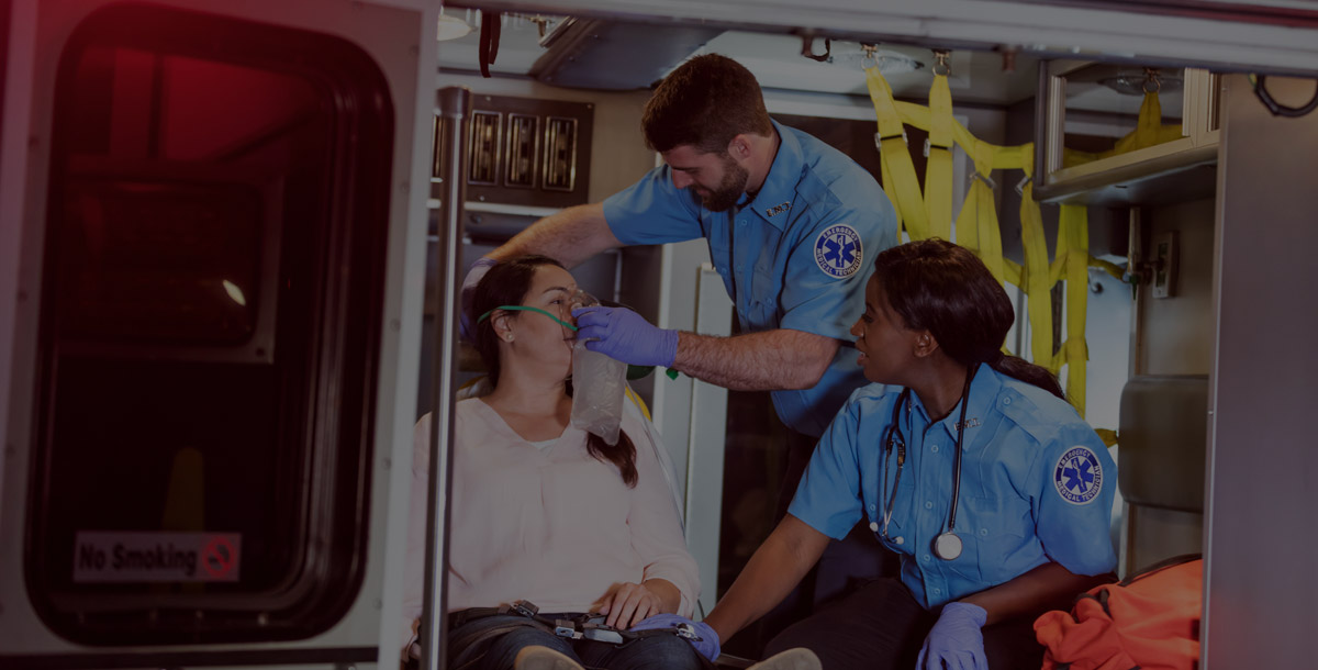 Two multi-ethnic paramedics, a Caucasian man and African-American woman, assisting a patient, giving her oxygen inside an ambulance. The patient, a mature Hispanic woman, looks worried.
