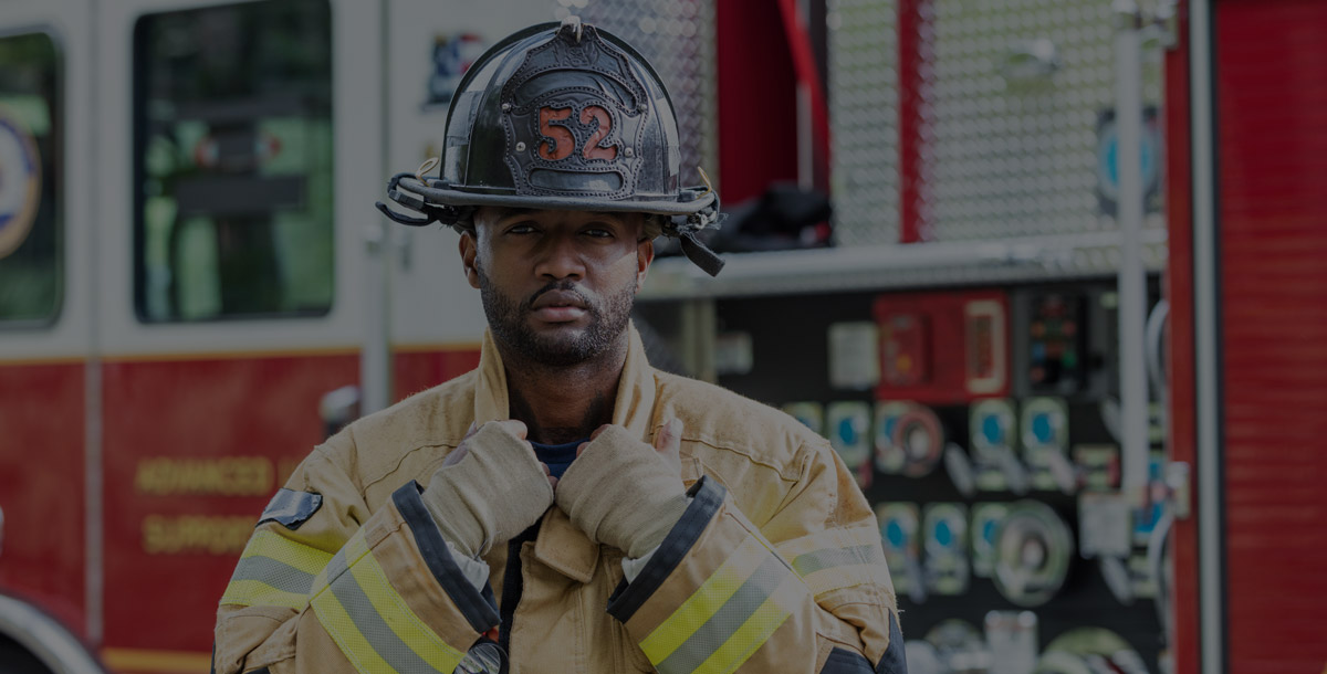 A firefighter standing in front of a fire truck, wearing a fire protection suit and helmet. He is a young African-American man in his 20s.