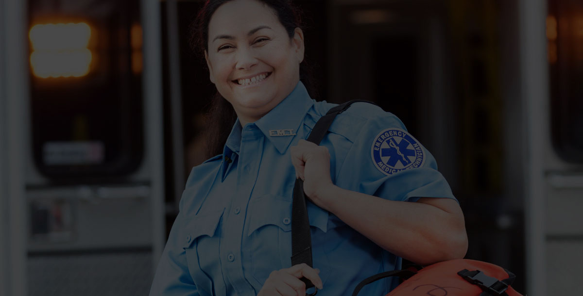 A mature Hispanic woman in her 40s, a paramedic, standing at the rear of an ambulance, by the open doors. She is looking at the camera with a confident expression, smiling, carrying a medical trauma bag on her shoulder.